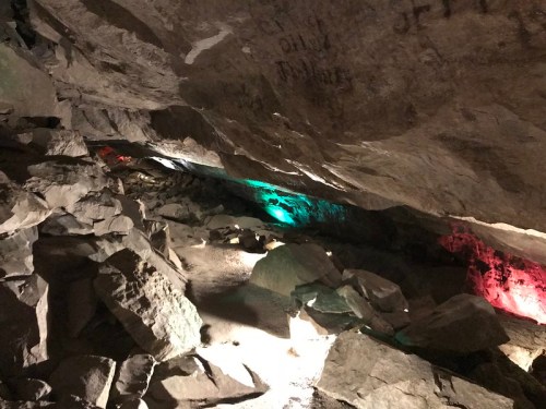 A dimly lit cave with colorful lights illuminating the rocky interior and scattered boulders on the ground.