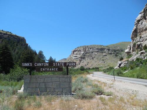 Sign for Sinks Canyon State Park at the entrance, surrounded by rocky hills and greenery under a clear blue sky.