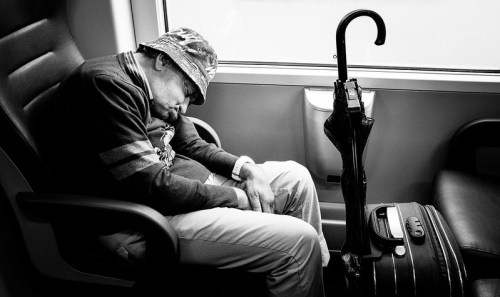 An elderly man in a hat sleeps on a train seat, with an umbrella and suitcase beside him. Black and white photo.