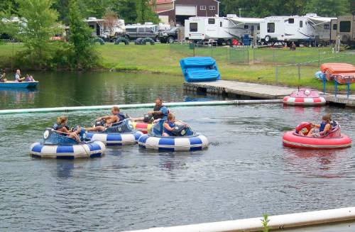 People enjoying bumper boats on a lake, with RVs and colorful inflatable rides in the background.