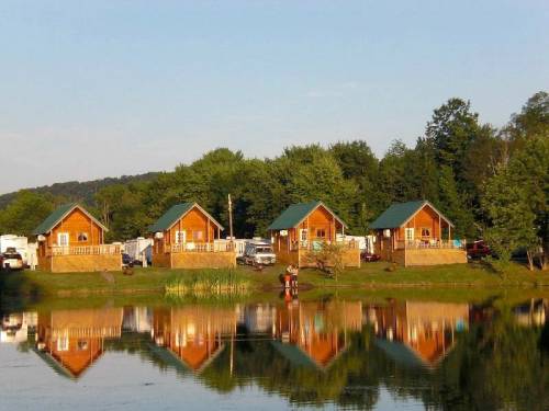 Four wooden cabins with green roofs reflect in a calm lake, surrounded by trees and a clear blue sky.