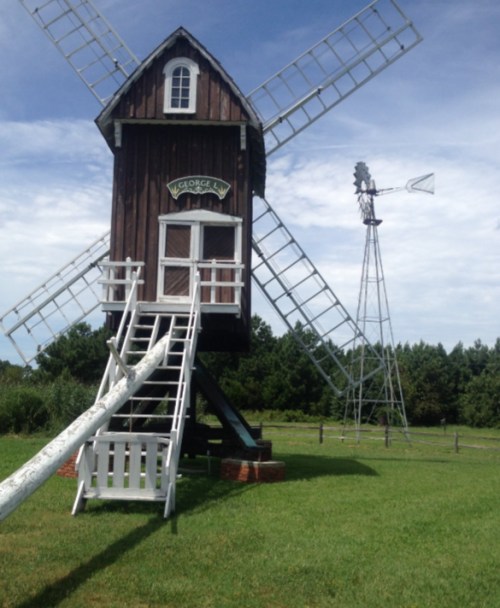 A wooden windmill with a staircase, surrounded by green grass and trees, with a wind pump nearby under a blue sky.