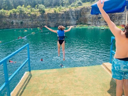 A child in a life jacket jumps off a platform into a blue swimming area, surrounded by people enjoying the water.