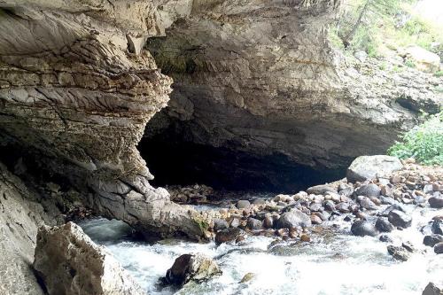 A rocky cave entrance beside a flowing river, surrounded by boulders and greenery.