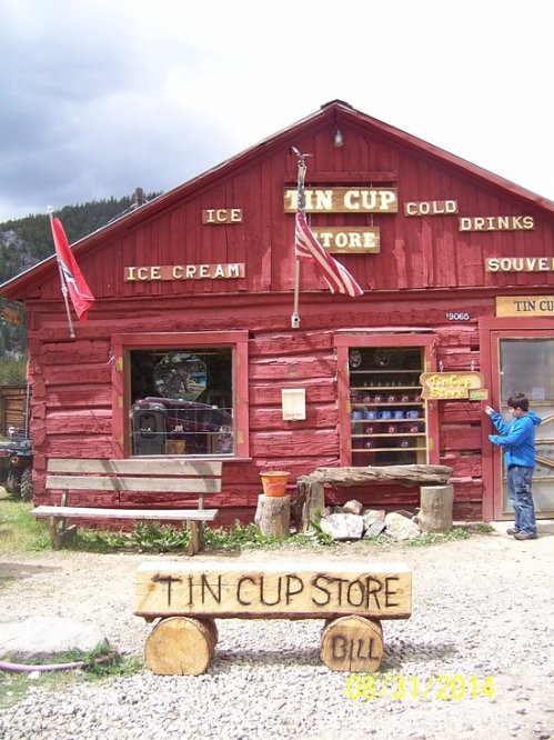 A rustic red wooden store with signs for ice cream and souvenirs, featuring a log bench and a person browsing outside.