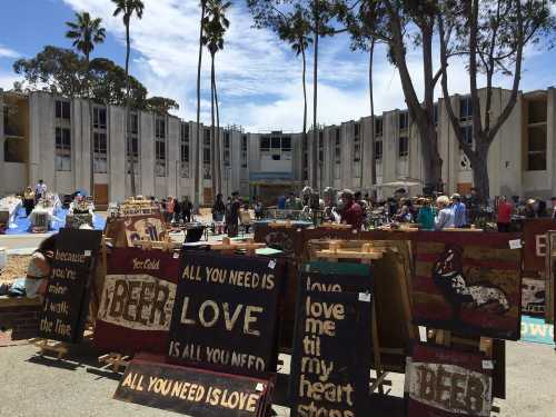 A bustling outdoor market with wooden signs displaying messages about love and beer, surrounded by palm trees and buildings.