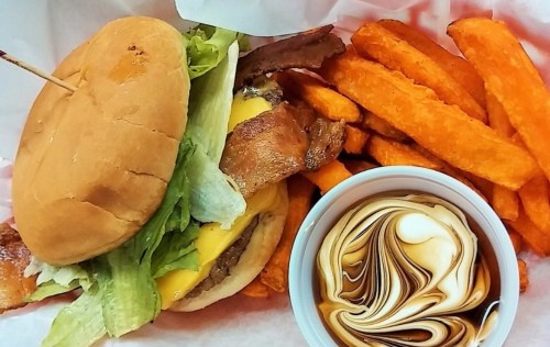A cheeseburger with lettuce and bacon, served with sweet potato fries and a dipping sauce in a small cup.