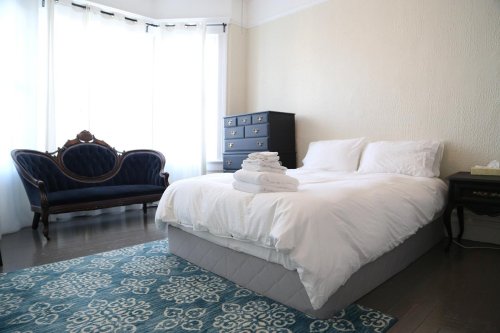 A bright bedroom featuring a large bed with white linens, a blue sofa, and a dark dresser against a light wall.