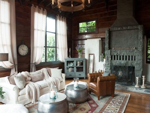 Cozy living room with wooden walls, a stone fireplace, plush furniture, and large windows letting in natural light.