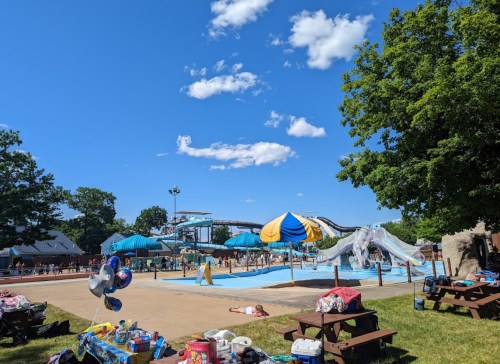 A sunny water park scene with slides, a pool, and people enjoying the day, surrounded by trees and picnic areas.