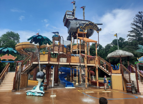 A colorful water play structure with slides, water features, and a pirate ship theme, surrounded by trees and blue sky.