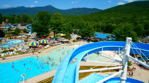 A vibrant water park scene with slides, a pool, and visitors enjoying the sunny day amidst green mountains.