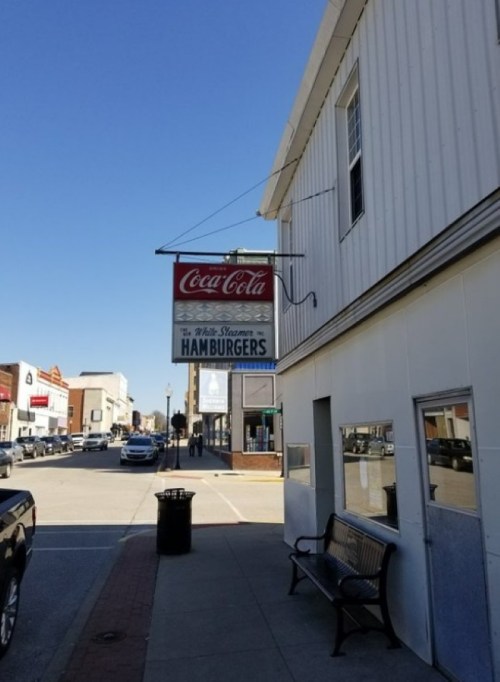 Sign for "Coca-Cola" and "White Steamer Hamburgers" on a building in a small town, with a clear blue sky above.