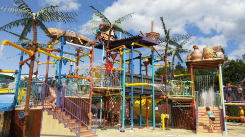 A colorful water play area with slides, fountains, and palm trees, filled with children enjoying the splash zone.