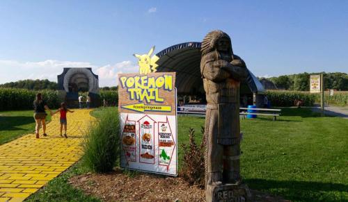 A colorful sign for "Pokemon Trail" near a wooden statue, with a yellow brick path leading into a cornfield.