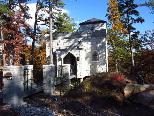 A small stone building surrounded by trees, featuring gothic-style architecture and a decorative fence.