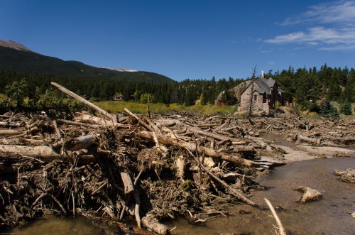 A stone house near a riverbank, surrounded by fallen trees and debris, with mountains and blue sky in the background.