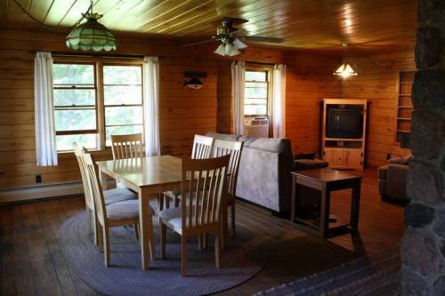Cozy wooden living room with a dining table, chairs, sofa, and TV, featuring large windows and warm lighting.