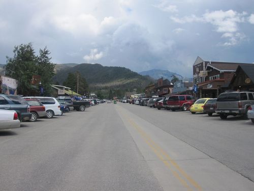 A wide street lined with parked cars and shops, set against a backdrop of mountains and cloudy skies.