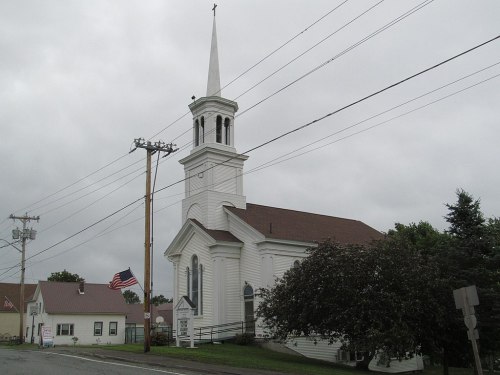 A white church with a tall steeple, surrounded by trees and power lines, under a cloudy sky. An American flag is visible.