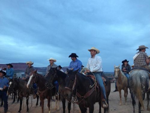 A group of cowboys on horseback, wearing hats and western attire, gathered outdoors against a cloudy sky.
