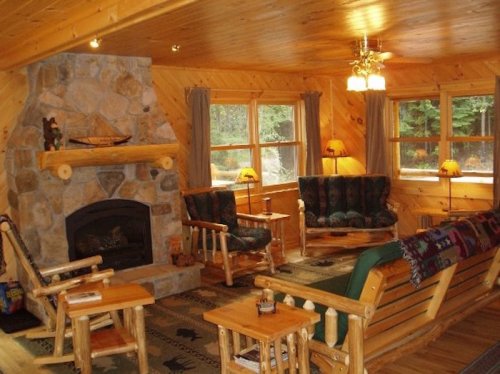 Cozy cabin interior with wooden walls, stone fireplace, comfortable seating, and large windows letting in natural light.