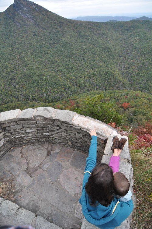A person with a child on their back points towards a distant mountain view from a stone lookout.