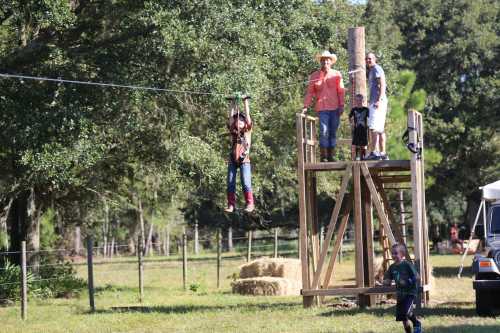 A group of people enjoy a zip line in a grassy area, with trees and hay bales in the background.