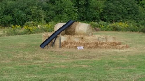 A black slide rests on a stack of hay bales in a grassy field, surrounded by greenery.