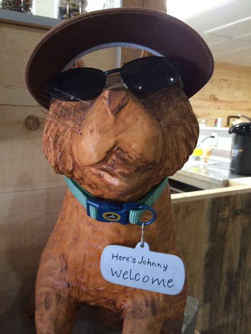 A wooden dog statue wearing sunglasses and a hat, with a sign that says "Here's Johnny, Welcome."