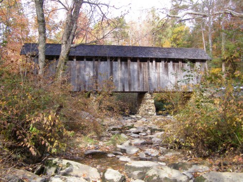 A wooden covered bridge spans a rocky stream, surrounded by autumn foliage and trees.