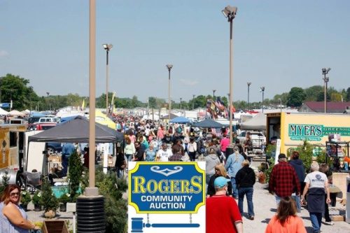 A bustling outdoor auction with crowds, tents, and vendors under a clear blue sky. "Rogers Community Auction" sign in foreground.