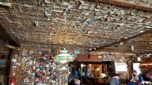 A rustic bar interior with a ceiling covered in dollar bills and walls adorned with stickers and memorabilia.