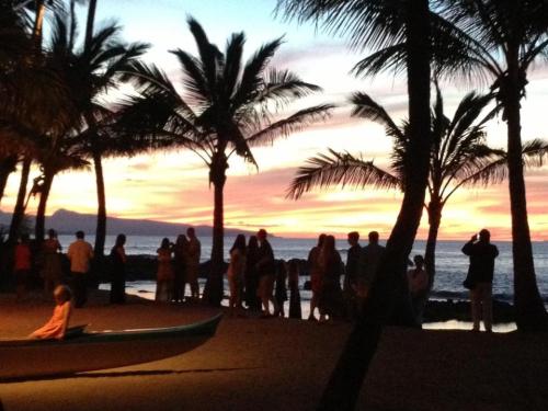 Silhouetted figures gather on a beach at sunset, with palm trees framing a colorful sky over the ocean.