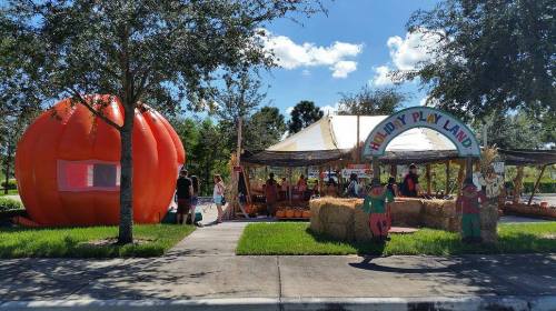 A vibrant pumpkin-shaped structure next to a festive play area with hay bales and colorful decorations under a blue sky.