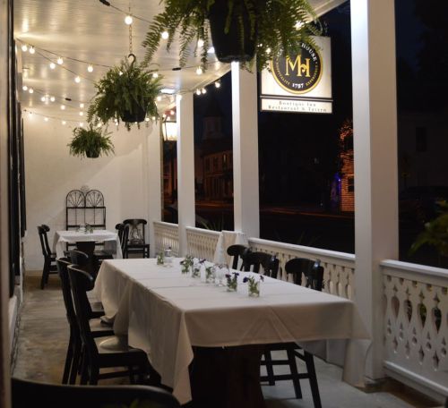 A dimly lit outdoor dining area with tables set for guests, surrounded by hanging plants and string lights.