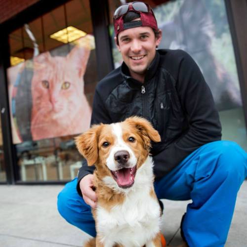 A man in a black jacket and blue pants smiles while sitting with a happy dog outside a pet store.