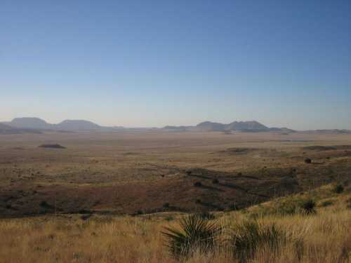 A vast, open landscape with rolling hills and mountains under a clear blue sky, featuring dry grass and sparse vegetation.