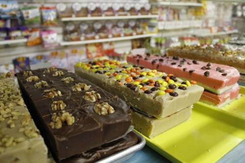 Colorful fudge bars with nuts and candy toppings displayed in a shop, with various snacks in the background.