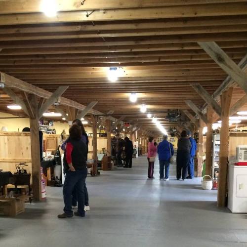 A spacious indoor market with wooden beams, showcasing various stalls and people browsing antiques and collectibles.