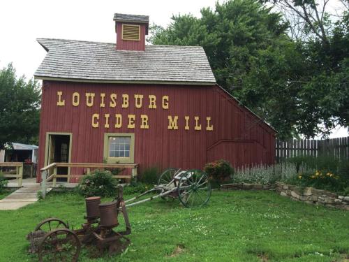 Red barn with "Louisburg Cider Mill" sign, surrounded by greenery and an old plow in the foreground.