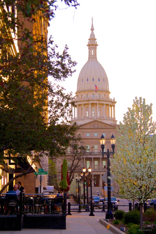 A view of a state capitol building with a dome, framed by trees and outdoor seating in the foreground during sunset.