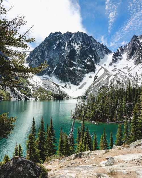 A serene mountain landscape featuring a turquoise lake, towering peaks, and lush evergreen trees under a blue sky.