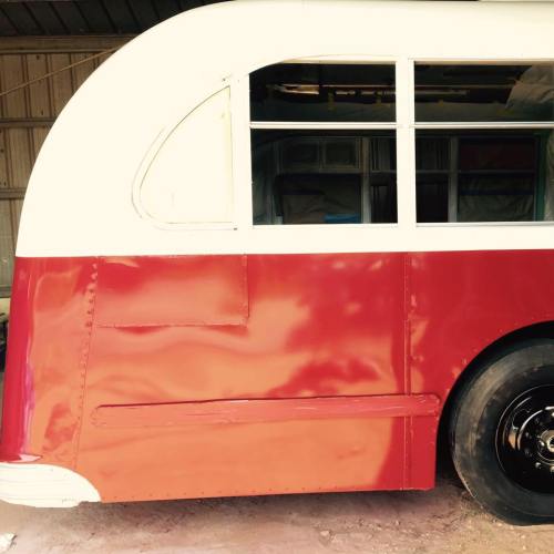 A vintage bus with a curved roof and red body, parked in a shed with a partially visible wheel.