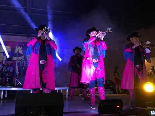 A lively band in colorful outfits performs on stage, featuring trumpet players and a drummer in the background.