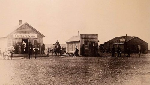 Historic black-and-white photo of a farmer's store with people and horses outside, set in a rural landscape.