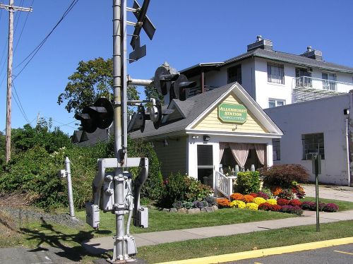 A quaint train station with a flower garden, next to a railroad crossing signal and a residential building in the background.