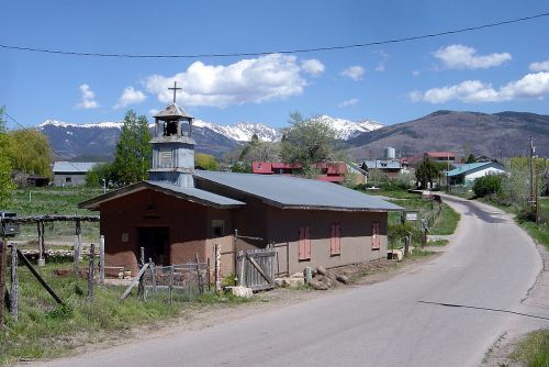A small church with a bell tower sits by a rural road, surrounded by mountains and a few buildings under a blue sky.