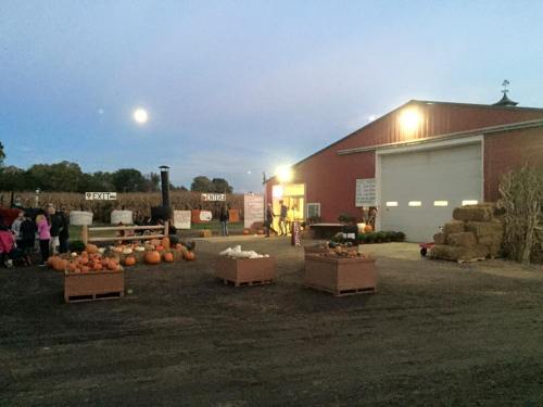 A farm scene at dusk with pumpkins, hay bales, and a red barn under a full moon. Children are gathered nearby.