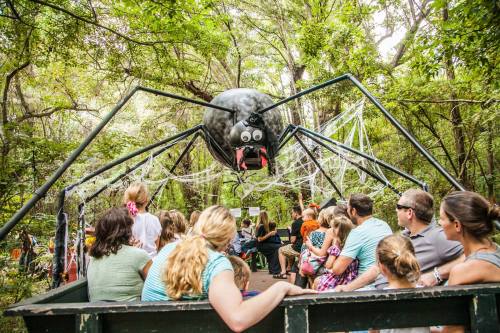 A large spider sculpture hangs over a group of people on a wagon ride through a wooded area decorated for Halloween.